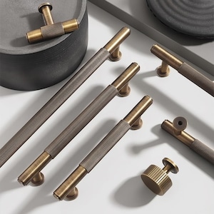 May include: Bronze-toned cabinet hardware, including handles and knobs, are arranged on a white surface. The handles have a ribbed design on the main bar and a smooth, rounded base. The knobs have a similar ribbed design.