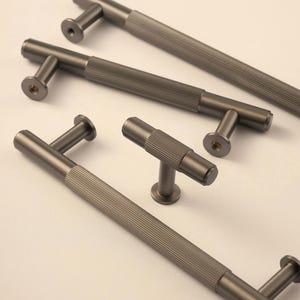 Pode incluir: Várias puxadores de armário cinza chumbo com um design texturizado e canelado. Os puxadores têm formato de T com uma alça cilíndrica e uma base redonda. O hardware está disposto em um fundo claro.