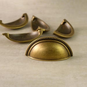 May include: A collection of antique brass-colored drawer pulls. The pulls are in a half-moon shape with a beaded edge. The hardware has a vintage aesthetic, suitable for furniture restoration or adding a classic touch to cabinets.