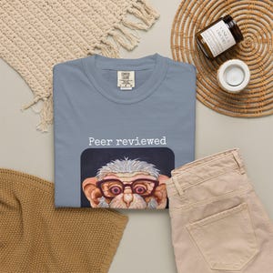 Può includere: Una felpa girocollo Comfort Colors azzurra con la scritta "Peer reviewed" sopra un'illustrazione a cartoni animati di una creatura con gli occhiali. La felpa è abbinata a pantaloni beige.