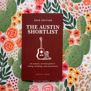 May include: A maroon book titled "THE AUSTIN SHORTLIST 2026 EDITION" with a guitar and cowboy boot graphic. The book is an annual guide to eating, drinking, and experiences in Austin, Texas. The background features a floral and cactus pattern.