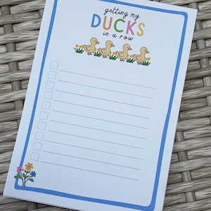 May include: A white rectangular notepad with a blue border featuring the text getting my DUCKS in a row in colorful letters. Below the text are four yellow ducklings walking in a line. The notepad includes ten blank lines with checkboxes.