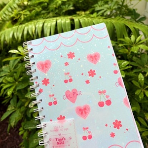 May include: A spiral-bound notebook with a light blue cover featuring a pattern of pink hearts, cherries, and flowers. The cover includes the text GRL PWR and a white label that reads 5.5 x 8.5 in, 60 sheets, 120 pages, That's So Savi.