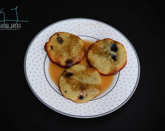 May include: Three golden brown pancakes with blueberries and a drizzle of syrup on a white plate with black polka dots.
