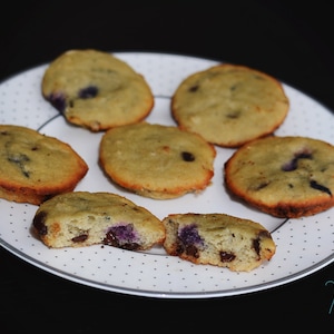 May include: A white plate with six small, round, light brown cookies with blueberries on top. The cookies are arranged in a circle on the plate. The plate is on a black surface.