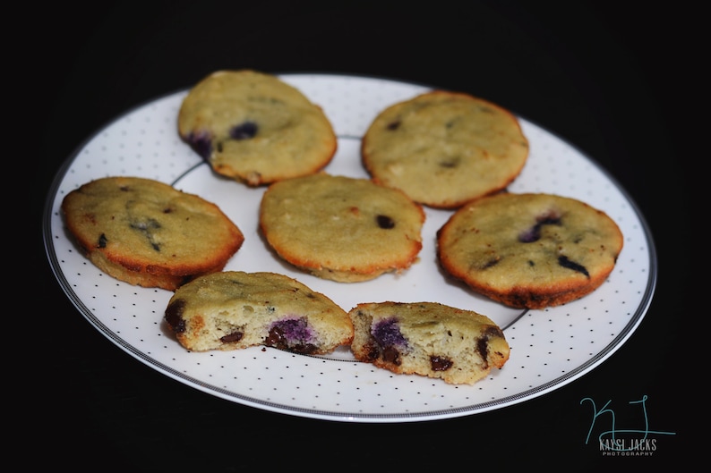 May include: A white plate with six small, round, light brown cookies with blueberries on top. The cookies are arranged in a circle on the plate. The plate is on a black surface.