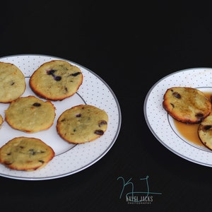 May include: Two white plates with a light brown background and white polka dots. One plate has six small, round, light brown cookies with dark blue blueberries. The other plate has three cookies with a light brown glaze and a dark brown sauce.