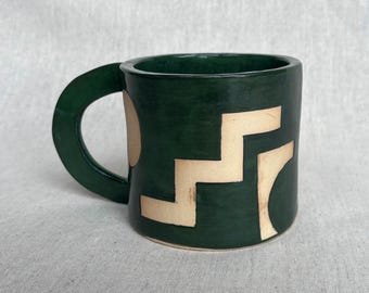 May include: A dark green ceramic mug featuring a large, curved handle and a light beige geometric pattern on the exterior. The design includes a stepped, staircase-like shape alongside a partial circular arc, creating a contrast against the dark glaze.