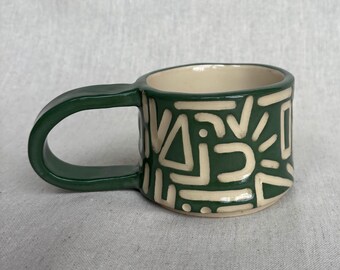 May include: A ceramic mug with a dark green glazed exterior featuring a cream-colored geometric pattern of lines, triangles, and circles. The mug has a wide, thick handle and a smooth, light-colored interior, showcasing a hand-crafted aesthetic.