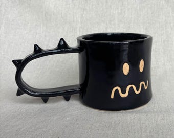 May include: A glossy black ceramic mug featuring a handle adorned with several pointed spikes. The front of the mug displays a hand-painted face consisting of two simple oval eyes and a wavy, horizontal line for a mouth, all in a light tan color.