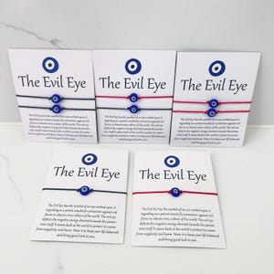 May include: Five white display cards labelled The Evil Eye, each featuring a blue and white eye symbol. The cards hold thin string bracelets in black or red, each adorned with a small blue, white, and black glass evil eye bead charm.