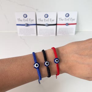 May include: Three adjustable cord bracelets in blue, black, and red, each featuring a central glass evil eye bead. The bracelets are displayed on white cards labelled The Evil Eye, which include descriptive text about the product's symbolism.