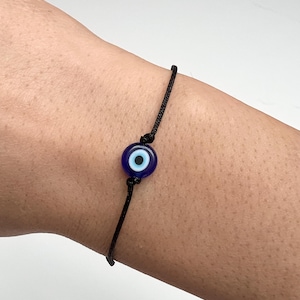 May include: A black cord bracelet with a blue and white evil eye charm. The charm is a round glass bead with a dark blue background and a white and black eye design. The bracelet is a simple, minimalist design.