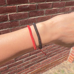 May include: Two braided cord bracelets worn on the wrist, one in a vibrant red colour and the other in a deep black colour. Both bracelets feature a woven texture and are constructed from thin, durable cord material with adjustable ends.