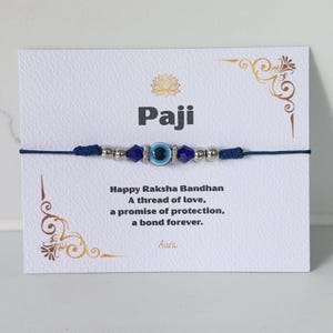 May include: A dark blue string bracelet featuring a central evil eye bead flanked by silver-toned metallic beads and dark blue faceted crystals. The bracelet is mounted on a white display card with the text Paji and Happy Raksha Bandhan.