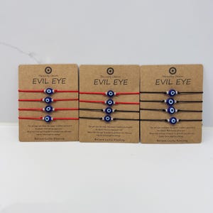 May include: Three brown cardboard display cards, each holding four thin string bracelets. The bracelets feature a small blue, white, and black evil eye bead. The cards are printed with the text "The Lucky Charm EVIL EYE" and "Believe-Lucky Blessing".