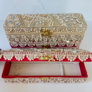 May include: Two rectangular jewellery boxes with gold-tone metal latches. The exterior features intricate gold embroidery, small white faux pearls, and reflective mirror accents. The open box reveals a cream-coloured interior lining and red fabric edges.