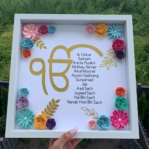 May include: A white shadow box frame containing a gold glitter Ik Onkar symbol and the Mool Mantar text in black. The display is decorated with various colourful paper flowers in shades of pink, blue, yellow, orange, and purple, along with gold glitter leaf accents.