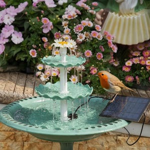 May include: A three-tiered, teal-colored garden fountain with water cascading from each level. A white column supports the tiers, topped with a daisy. A small bird perches on a solar panel next to the fountain, surrounded by flowers.