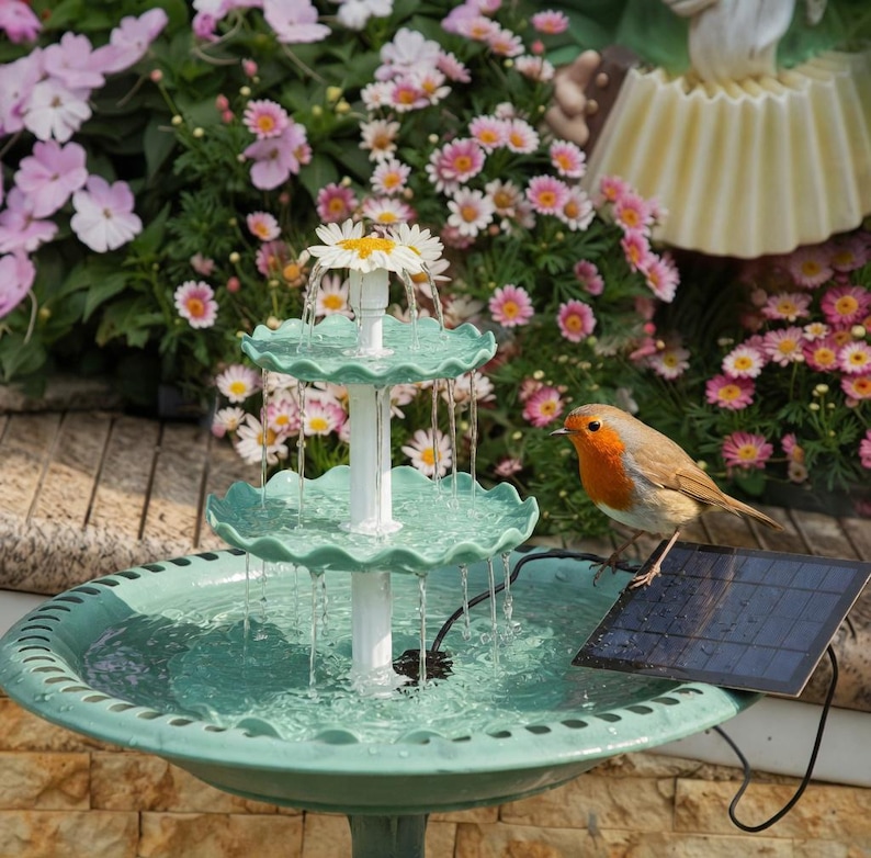 May include: A three-tiered, teal-colored garden fountain with water cascading from each level. A white column supports the tiers, topped with a daisy. A small bird perches on a solar panel next to the fountain, surrounded by flowers.