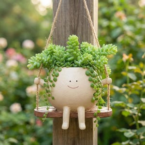 May include: A round, cream-coloured ceramic planter with a simple smiling face, two arms, and two legs. The planter sits on a small wooden swing suspended by jute twine, containing a vibrant green succulent plant with trailing stems and leaves.