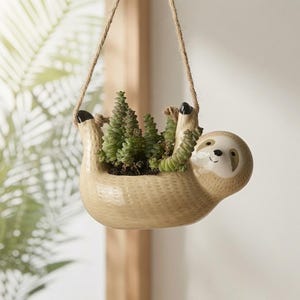 Può includere: Un vaso sospeso in ceramica beige a forma di bradipo con superficie testurizzata, un volto sorridente con macchie scure intorno agli occhi e artigli neri. Il vaso è appeso a una corda di iuta e contiene una pianta succulenta di colore verde.