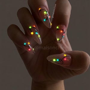 May include: Almond-shaped nails with a sheer nude base featuring a hand-painted black wire design adorned with small, glowing multicolored Christmas light bulbs in red, green, yellow, and blue. The text @maisonchiee is visible on the nails.