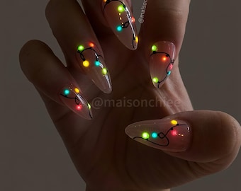 May include: Almond-shaped nails with a sheer nude base featuring a hand-painted black wire design adorned with small, glowing multicolored Christmas light bulbs in red, green, yellow, and blue. The text @maisonchiee is visible on the nails.