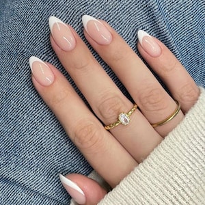 Op de afbeelding: Een hand met amandelvormige nagels met een klassieke Franse manicure met witte uiteinden. De nagels zijn gelakt met een transparante, natuurlijk ogende roze nagellak. Twee gouden ringen, waarvan één met een ovale, heldere steen.