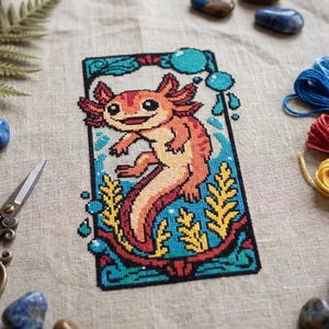 May include: A cross-stitch design featuring a smiling axolotl in shades of orange, red, and yellow, set against a blue and green aquatic background. The design is framed with a black border and surrounded by blue and yellow embroidery floss, scissors, and decorative stones.