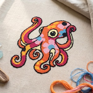 May include: A vibrant cross-stitch octopus design on a beige fabric. The octopus is rendered in orange, pink, yellow, and blue. The design is outlined in black, with detailed tentacles and eyes. Threads are visible.