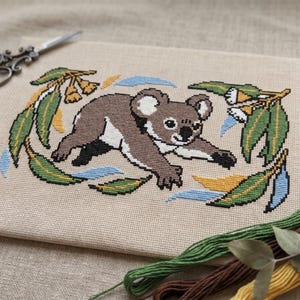 May include: Cross-stitch embroidery of a koala bear with eucalyptus leaves and branches. The koala is brown, white, and black, surrounded by green, blue, and yellow leaves. Scissors and embroidery floss are in the background.