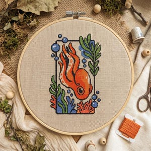 Peut inclure: Un cercle de broderie au point de croix avec un motif de poulpe orange, des bulles bleues et des algues vertes. La pieuvre est encadrée dans un rectangle noir. Le fond est un tissu beige, et le cercle est en bois.