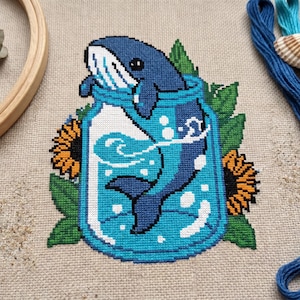 May include: Cross-stitch design of a blue whale in a jar with water and bubbles, surrounded by green leaves and orange sunflowers. Set against a tan fabric background.