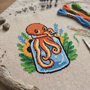May include: Cross-stitch art featuring an orange octopus inside a blue jar, surrounded by colorful flowers and green leaves. The artwork is on a beige fabric, with embroidery floss in orange, blue, and green in the background.
