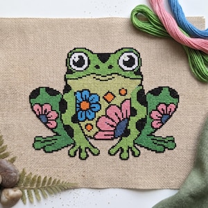 May include: A cross-stitch frog design on tan fabric. The frog is green with black spots and floral accents in pink, blue, and orange. Embroidery floss in green, pink, and blue are visible in the background. The craft project is on a white surface.