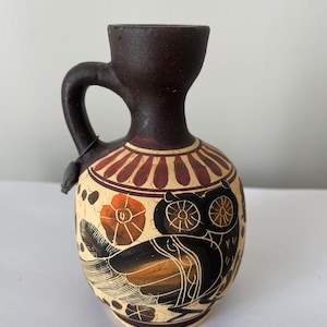Puede incluir: Un pequeño jarrón de cerámica pintado a mano con asa y borde marrón oscuro. El jarrón presenta un cuerpo color crema con un diseño de búho negro y naranja, junto con patrones florales y geométricos. El jarrón mide unos 13 cm de alto.