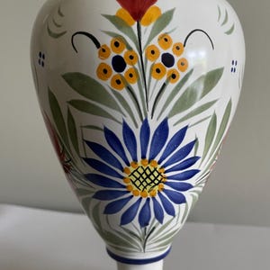 Pode incluir: Um vaso de cerâmica branco com desenhos florais pintados à mão. O vaso apresenta uma grande flor azul, detalhes em amarelo e laranja e folhas verdes. O vaso tem aproximadamente 25 cm de altura.