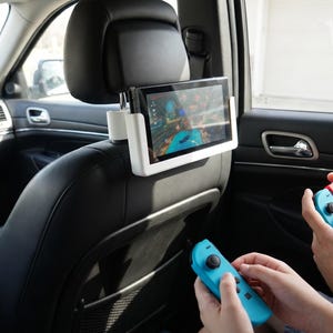 Può includere: Un supporto bianco per tablet, che mostra un videogioco, è attaccato al poggiatesta di un'auto. Due persone tengono in mano controller di gioco blu e rossi. L'interno dell'auto è visibile.