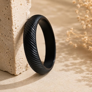 May include: A black ring with a textured, ridged design. The ring is leaning against a beige, porous stone. Dried flowers are in the background, and the ring is casting a shadow on the surface.
