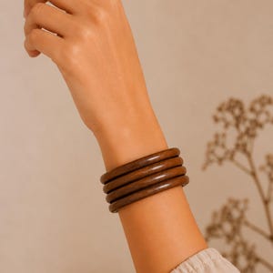 May include: A set of five dark brown wooden bangles are stacked on a wrist. The bangles are smooth and have a polished finish. The background is a neutral beige color, with a hint of dried flowers.