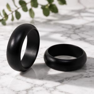 May include: Two solid black rings with a smooth, matte finish and a rounded, domed profile. One ring is positioned upright, while the other lies flat on its side. The rings feature a simple, continuous band design with no visible engravings or stones.