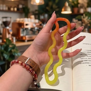 May include: A wavy, flexible, orange and yellow toy held in a hand. The toy is a vibrant gradient of colours. A brown leather watch with a silver buckle and a beaded bracelet are also visible.