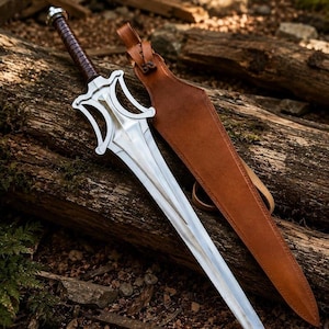 May include: A silver-coloured metal sword with a complex, curved crossguard and a dark brown leather-wrapped handle. Beside the sword lies a matching tan leather scabbard featuring a pointed shape and a leather strap attachment at the top.