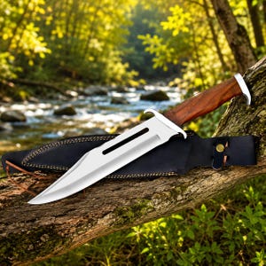 May include: A fixed-blade knife with a polished silver steel blade featuring a long central fuller and a dark brown wooden handle. The knife rests on a black leather sheath with visible stitching and a single brass-coloured metal snap button.