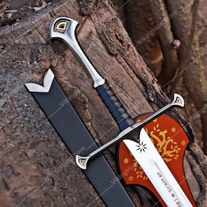 May include: A silver-toned metal sword with a black leather-wrapped grip and a pommel featuring a gold-coloured triangular inlay. The blade displays an engraved starburst pattern and runic text. A black scabbard with a silver tip is positioned nearby.