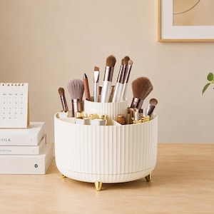 May include: A cream-coloured, cylindrical rotating makeup brush organiser with vertical ribbed detailing and three small gold-toned feet. The organiser features multiple interior compartments filled with various makeup brushes and cosmetic tools.