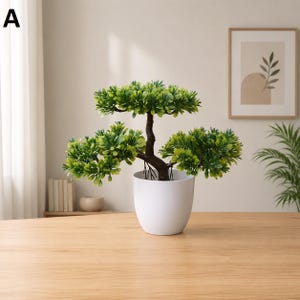 May include: An artificial bonsai tree in a white ceramic pot. The tree features a brown trunk and branches, adorned with bright green leaves. The pot is round and rests on a light-coloured wooden surface. The background includes a framed picture and a glimpse of a curtain.