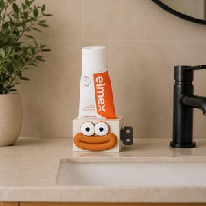 Puede incluir: Un tubo blanco de pasta de dientes elmex con una franja naranja se coloca en un dispensador de pasta de dientes novedoso. El dispensador es un cubo beige con ojos y boca de dibujos animados. Un grifo negro y una planta están en el fondo.
