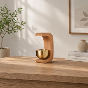 May include: A wooden desk bell with a curved wooden frame and a gold-coloured bell. A small brass ball hangs inside the bell. The bell is on a wooden table with a plant and books in the background.